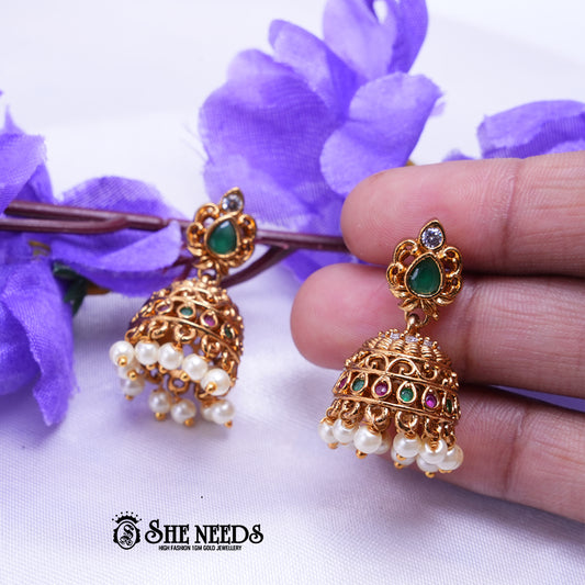 Gold earrings with green held by a hand against a purple flower background, with 'SHE NEEDS' branding.
