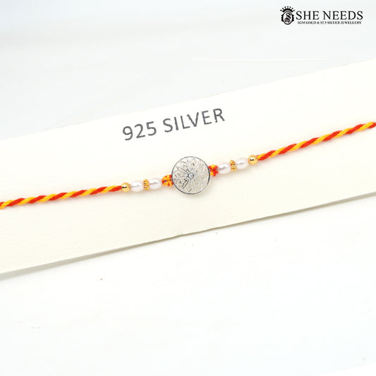 A 925 sterling silver Rakhi bracelet with a central engraved silver disc, flanked by white pearl beads and small gold spacer beads, strung on a red and yellow braided cord. The bracelet is displayed on a white card labeled '925 SILVER' with the brand logo 'SHE NEEDS' in the top right corner.
