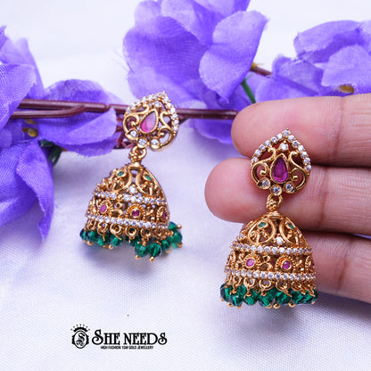 Antique gold Aaradhya jhumka earrings featuring sparkling CZ stones, ruby accents, intricate filigree detailing, and emerald green bead drops.|She Needs Jewellery
