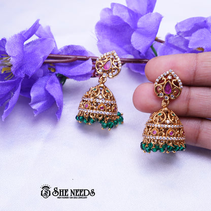 Antique gold Aaradhya jhumka earrings featuring sparkling CZ stones, ruby accents, intricate filigree detailing, and emerald green bead drops.|She Needs Jewellery
