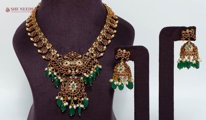 Shop the Antique Green Beaded Temple Necklace Set with matching jhumka earrings. Crafted with ruby stones, green beads, pearls, and antique gold polish, perfect for bridal, festive, and traditional occasions.
