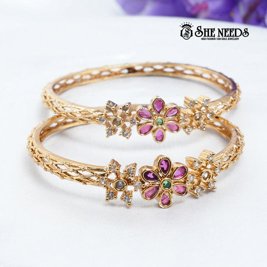 Gold-plated floral designer bangles pair featuring ruby-pink kemp stones, green center stone, CZ embellishments, and intricate openwork craftsmanship on a white background |She Needs Jewellery