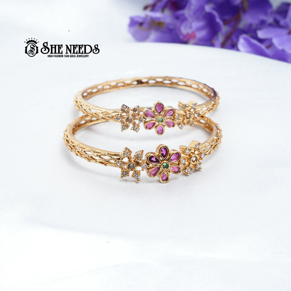 Gold-plated floral designer bangles pair featuring ruby-pink kemp stones, green center stone, CZ embellishments, and intricate openwork craftsmanship on a white background |She Needs Jewellery