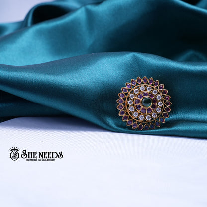 Antique gold circular saree pin featuring concentric layers of kundan stones, ruby-pink accents, and an emerald-green center stone in a radiant sunburst design.|She Needs Jewellery
