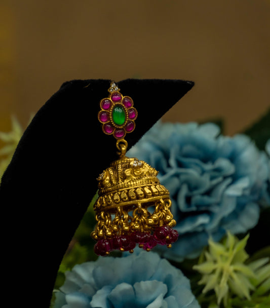 Gold jhumka earring with colorful stones on a black stand against a floral background