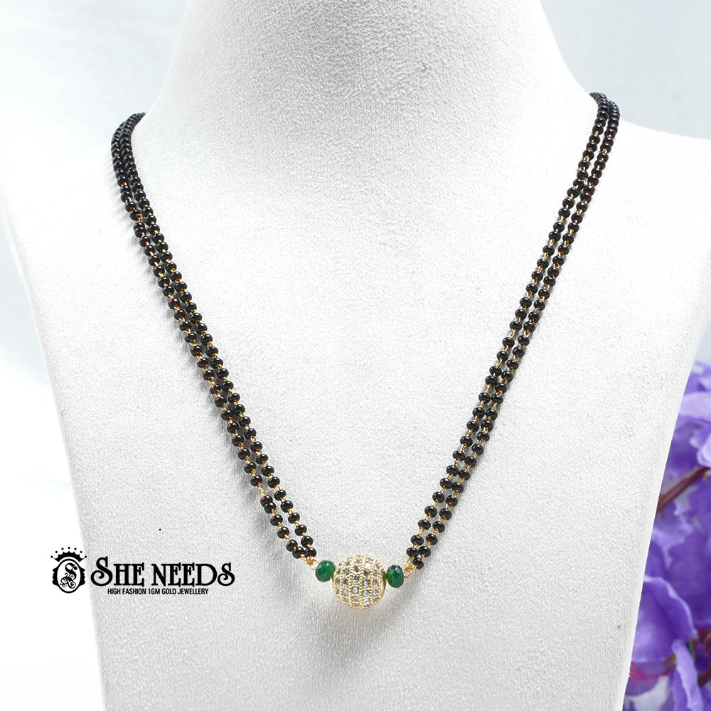 Emerald green dual-string mangalsutra with black beads, CZ-studded spherical pendant, green bead accents, and gold-tone finish displayed on a white jewelry bust |She Needs Jewellery
