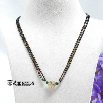Emerald green dual-string mangalsutra with black beads, CZ-studded spherical pendant, green bead accents, and gold-tone finish displayed on a white jewelry bust |She Needs Jewellery
