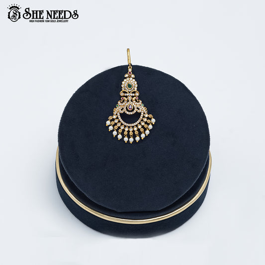 Antique gold Grand Chandbali Temple Tikka featuring sparkling kundan stones, emerald-green accents, intricate chandbali craftsmanship, and cascading pearl danglers displayed on a navy velvet stand.|She Needs Jewellery
