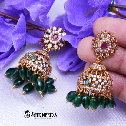 Antique gold Kavya jhumka earrings featuring sparkling CZ stones, ruby center accents, green stone details, and green bead drops.|She Needs Jewellery
