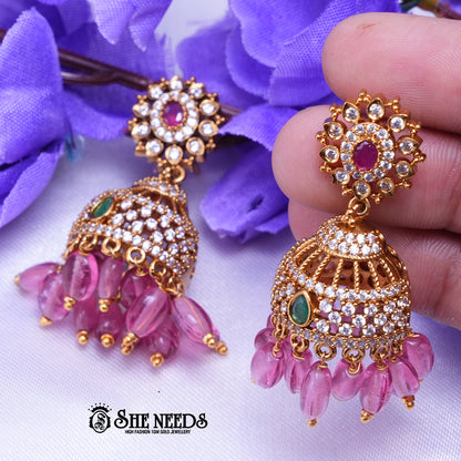 Antique gold Kavya jhumka earrings featuring sparkling CZ stones, ruby center accents, green stone details, and pink bead drops.|She Needs Jewellery

