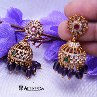 Antique gold Kavya jhumka earrings featuring sparkling CZ stones, ruby center accents, green stone details, and purple bead drops.|She Needs Jewellery
