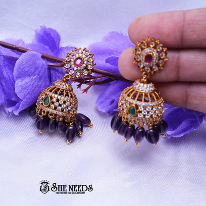 Antique gold Kavya jhumka earrings featuring sparkling CZ stones, ruby center accents, green stone details, and purple bead drops.|She Needs Jewellery
