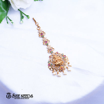 Antique gold Lakshmi Ruby Pearl Maang Tikka featuring a Goddess Lakshmi motif, sparkling kundan stones, ruby-pink and green accents, intricate temple jewelry detailing, and elegant pearl drop danglers.|She Needs Jewellery
