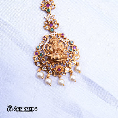 Antique gold Lakshmi Ruby Pearl Maang Tikka featuring a Goddess Lakshmi motif, sparkling kundan stones, ruby-pink and green accents, intricate temple jewelry detailing, and elegant pearl drop danglers.|She Needs Jewellery
