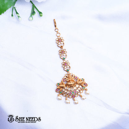 Antique gold Lakshmi Ruby Pearl Maang Tikka featuring a Goddess Lakshmi motif, sparkling kundan stones, ruby-pink and green accents, intricate temple jewelry detailing, and elegant pearl drop danglers.|She Needs Jewellery
