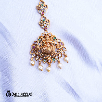 Antique gold Lakshmi Triad Pearl Maang Tikka featuring a Goddess Lakshmi motif, sparkling kundan stones, pink and green accents, intricate temple jewelry detailing, and dangling pearl drops on a white fabric background | She Needs Jewellery