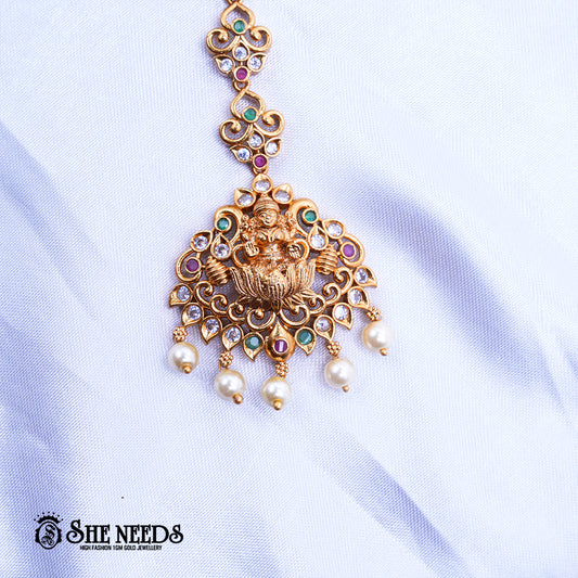 Antique gold Lakshmi Triad Pearl Maang Tikka featuring a Goddess Lakshmi motif, sparkling kundan stones, pink and green accents, intricate temple jewelry detailing, and dangling pearl drops on a white fabric background | She Needs Jewellery