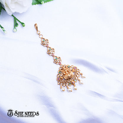 Antique gold Lakshmi Triad Pearl Maang Tikka featuring a Goddess Lakshmi motif, sparkling kundan stones, pink and green accents, intricate temple jewelry detailing, and dangling pearl drops on a white fabric background |She Needs Jewellery
