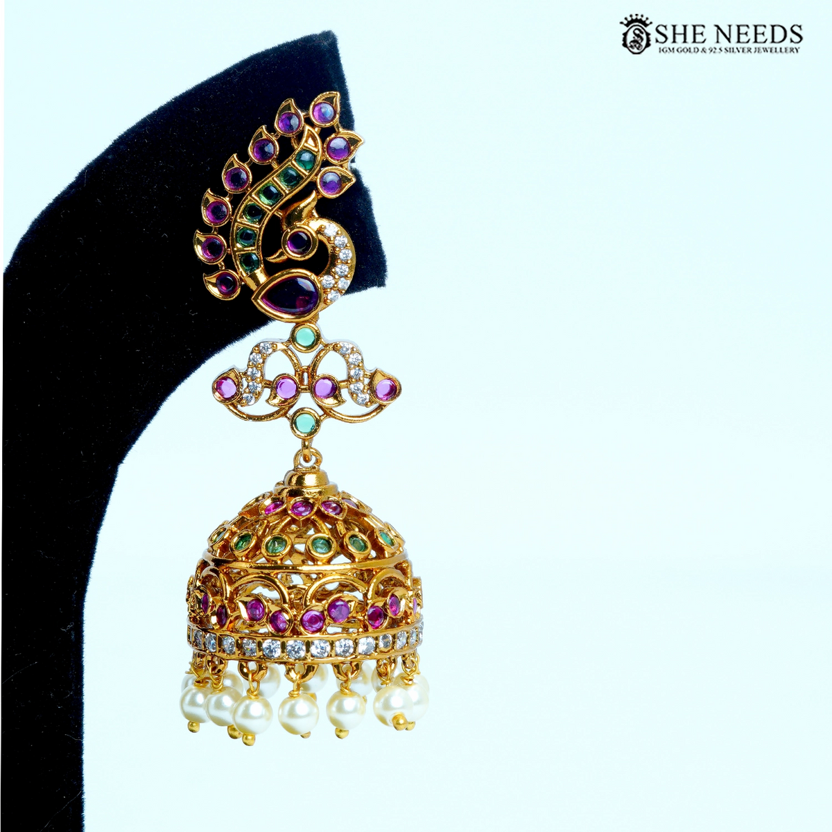 Pair of Mayura Royale peacock curve jhumka earrings with pink and green stones, white accents and pearl drop detailing on a black jewellery stand|She Needs Jewellery
