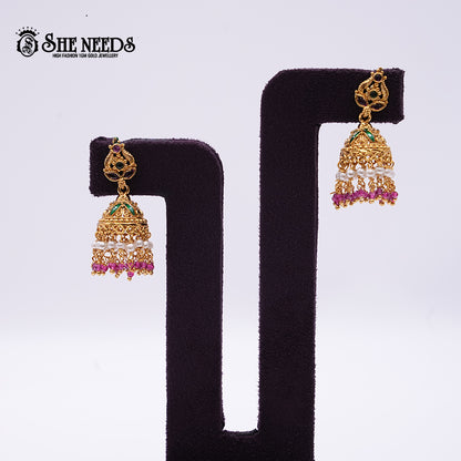 Antique gold Nakshi Pearl Cascade Jhumkas featuring emerald-green and pink stones, layered pearl danglers, pink bead drops, and intricate temple-style craftsmanship displayed on a velvet stand.|She Needs Jewellery
