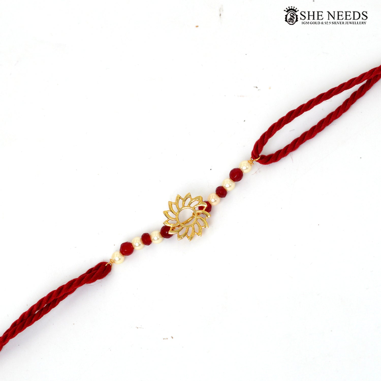 A red braided string bracelet with a central gold lotus flower charm and small red beads, shown on a plain white background. The brand logo 'SHE NEEDS' is visible in the top right corner.