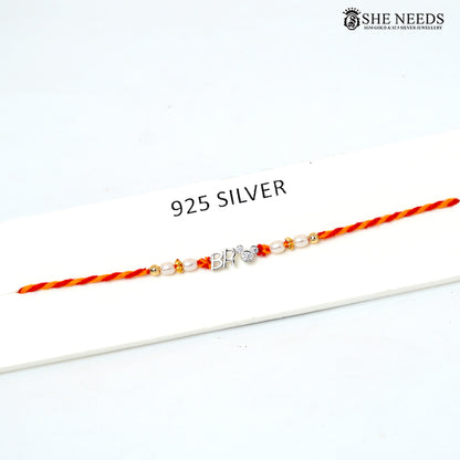A 925 silver Rakhi bracelet with a 'BRO' initial charm and small beads, displayed on a white card with the text '925 SILVER'. The brand logo 'SHE NEEDS' is visible in the top right corner.