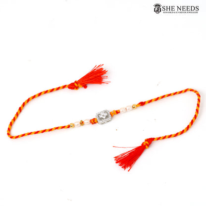 A traditional Indian Rakhi featuring a braided red and yellow cord, adorned with small white beads, a central silver-toned stone, and vibrant red tassels at both ends, displayed on a clean white background with a brand logo in the top right corner.