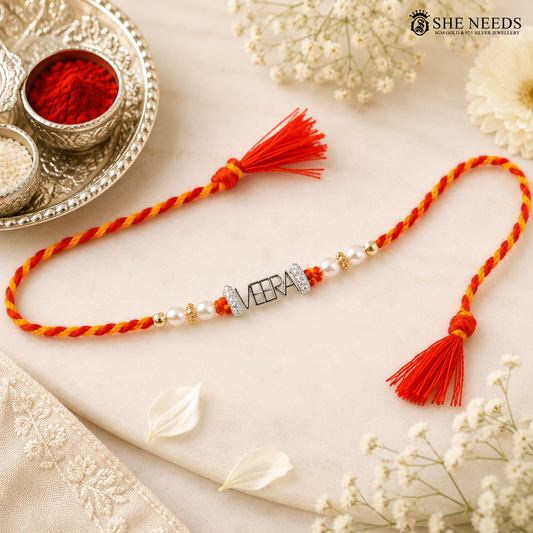 A braided Rakhi with red and yellow cords, white pearl beads, and a silver-tone 'VEERA' nameplate, laid on a cream fabric with decorative flowers and a silver tray in the background. SHE NEEDS logo is visible in the top right.