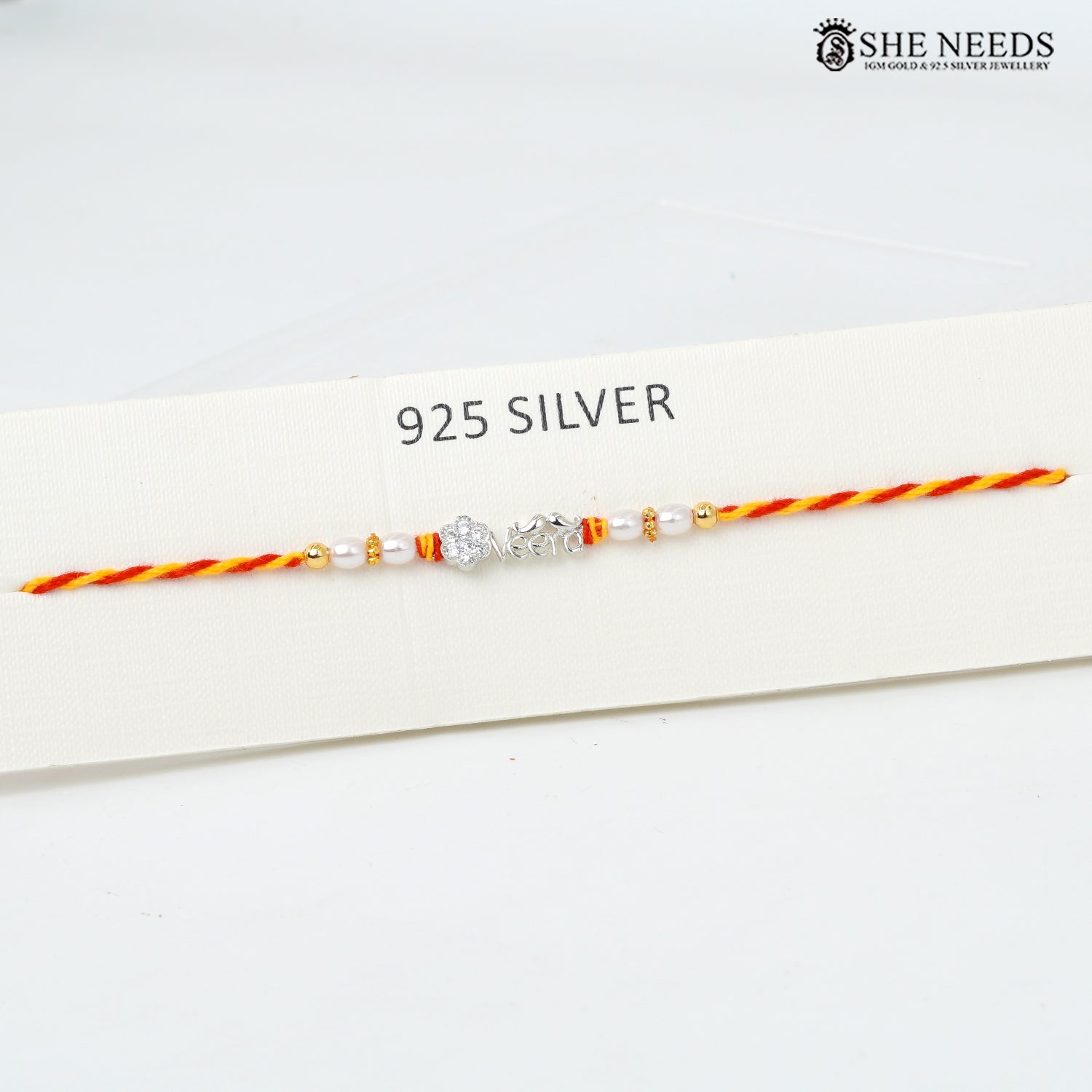 A delicate 925 sterling silver Rakhi bracelet with a silver VEERA charm, white pearl beads, and orange-red and yellow-orange braided cord, displayed on a white card labeled '925 SILVER' with the She Needs brand logo in the top right.
