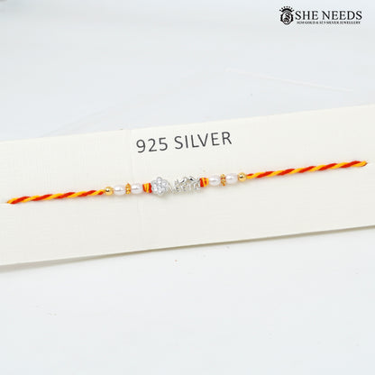 A delicate 925 sterling silver Rakhi bracelet with a silver VEERA charm, white pearl beads, and orange-red and yellow-orange braided cord, displayed on a white card labeled '925 SILVER' with the She Needs brand logo in the top right.