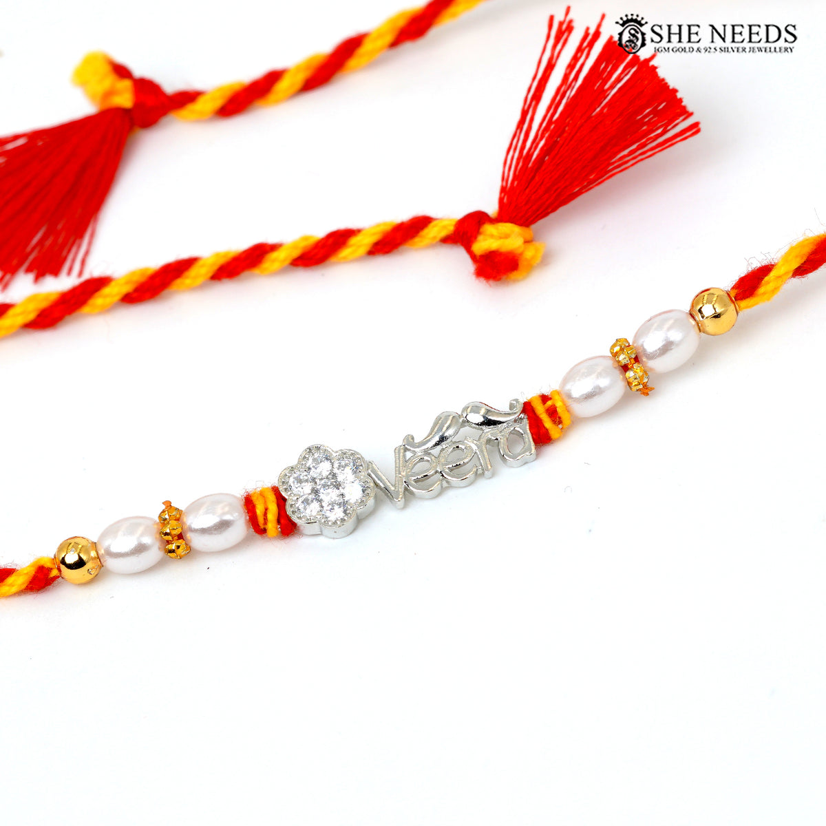 A close-up of a braided Rakhi bracelet with red and yellow cords, white faux pearls, gold spacer beads, and a silver nameplate engraved with 'Veera'. A second matching bracelet with tassels is partially visible in the background.