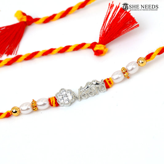 A close-up of a braided Rakhi bracelet with red and yellow cords, white faux pearls, gold spacer beads, and a silver nameplate engraved with 'Veera'. A second matching bracelet with tassels is partially visible in the background.
