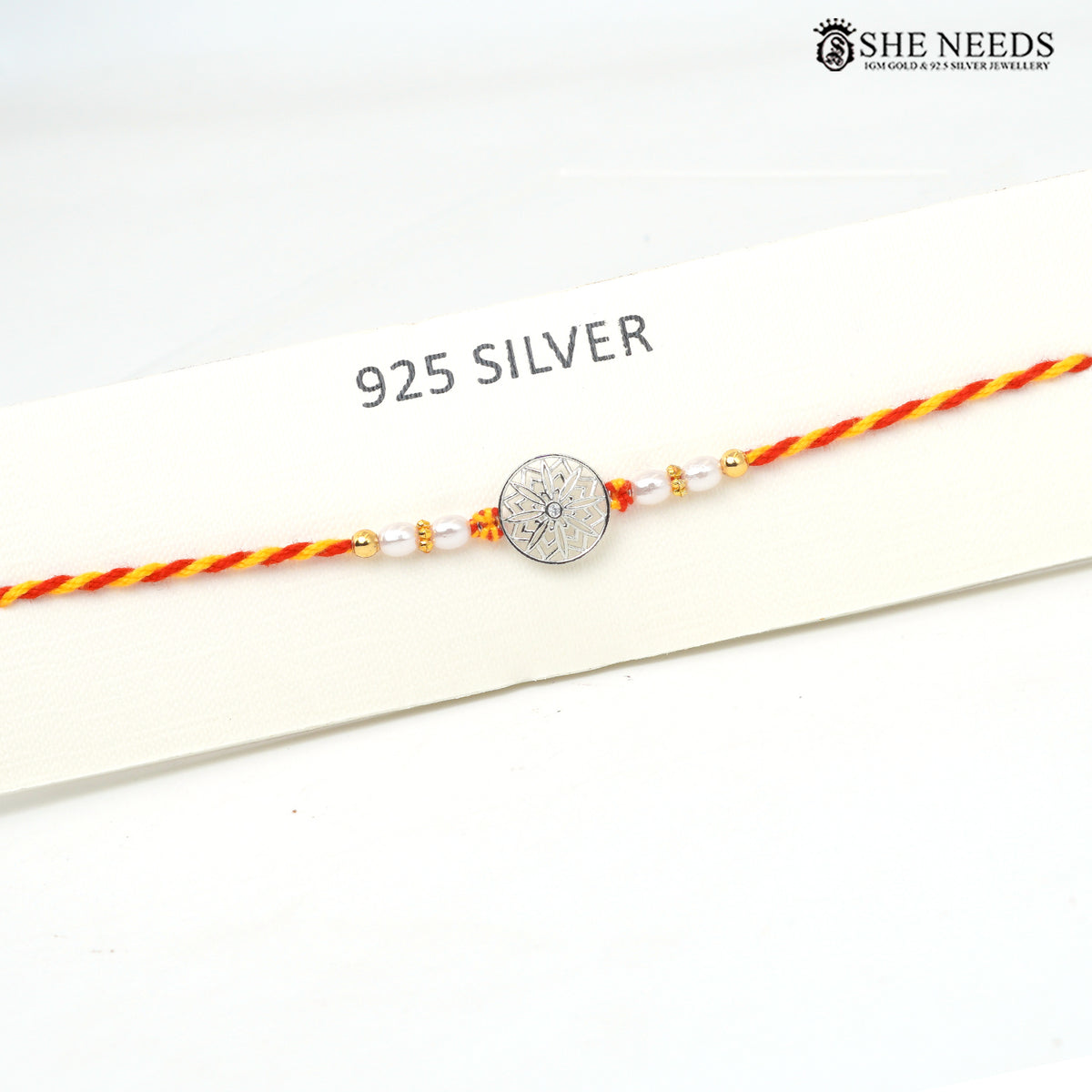 A 925 sterling silver Rakhi bracelet with a central engraved silver disc, flanked by white pearl beads and small gold spacer beads, strung on a red and yellow braided cord. The bracelet is displayed on a white card labeled '925 SILVER' with the brand logo 'SHE NEEDS' in the top right corner.