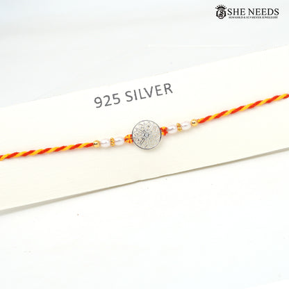 A 925 sterling silver Rakhi bracelet with a central engraved silver disc, flanked by white pearl beads and small gold spacer beads, strung on a red and yellow braided cord. The bracelet is displayed on a white card labeled '925 SILVER' with the brand logo 'SHE NEEDS' in the top right corner.
