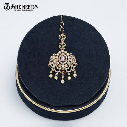 Antique gold Peacock Motif Temple Tikka featuring a pink teardrop stone, sparkling CZ stones, green accents, intricate peacock detailing, and pearl drop danglers displayed on a navy velvet stand |She Needs Jewellery