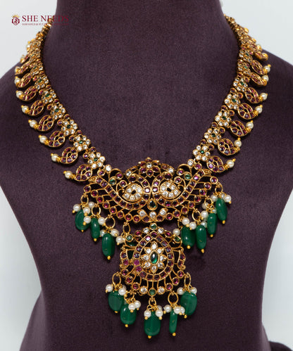 Shop the Antique Green Beaded Temple Necklace Set with matching jhumka earrings. Crafted with ruby stones, green beads, pearls, and antique gold polish, perfect for bridal, festive, and traditional occasions.