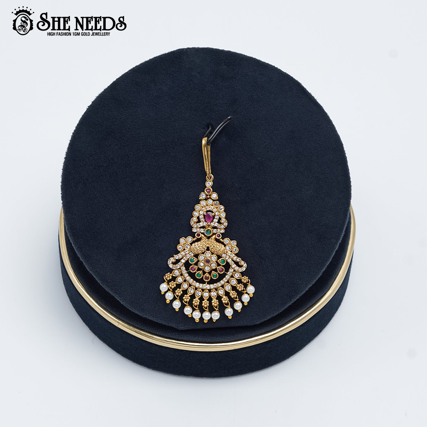 Antique gold Royal Chandbali Temple Tika featuring a goddess motif, sparkling kundan stones, ruby-pink and emerald-green accents, intricate chandbali detailing, and elegant pearl danglers displayed on a navy velvet stand.|She Needs Jewellery

