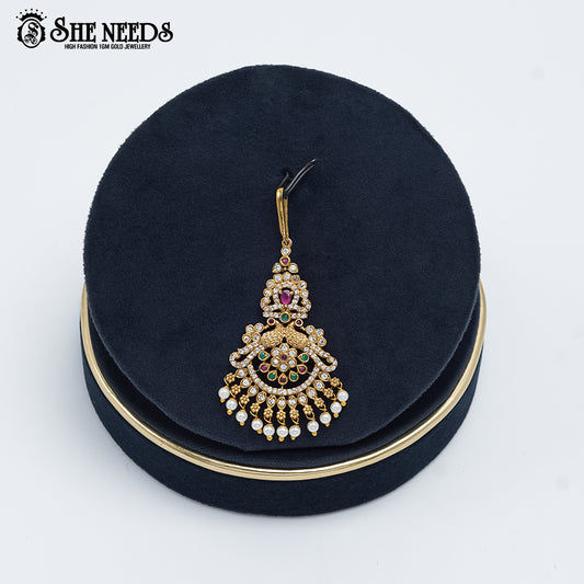 Antique gold Royal Chandbali Temple Tika featuring a goddess motif, sparkling kundan stones, ruby-pink and emerald-green accents, intricate chandbali detailing, and elegant pearl danglers displayed on a navy velvet stand.|She Needs Jewellery
