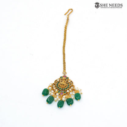 Traditional Lakshmi maang tikka with gold-toned detailing, emerald green stones, ruby accents, white stones and pearl drop embellishments|She Needs Jewellery
