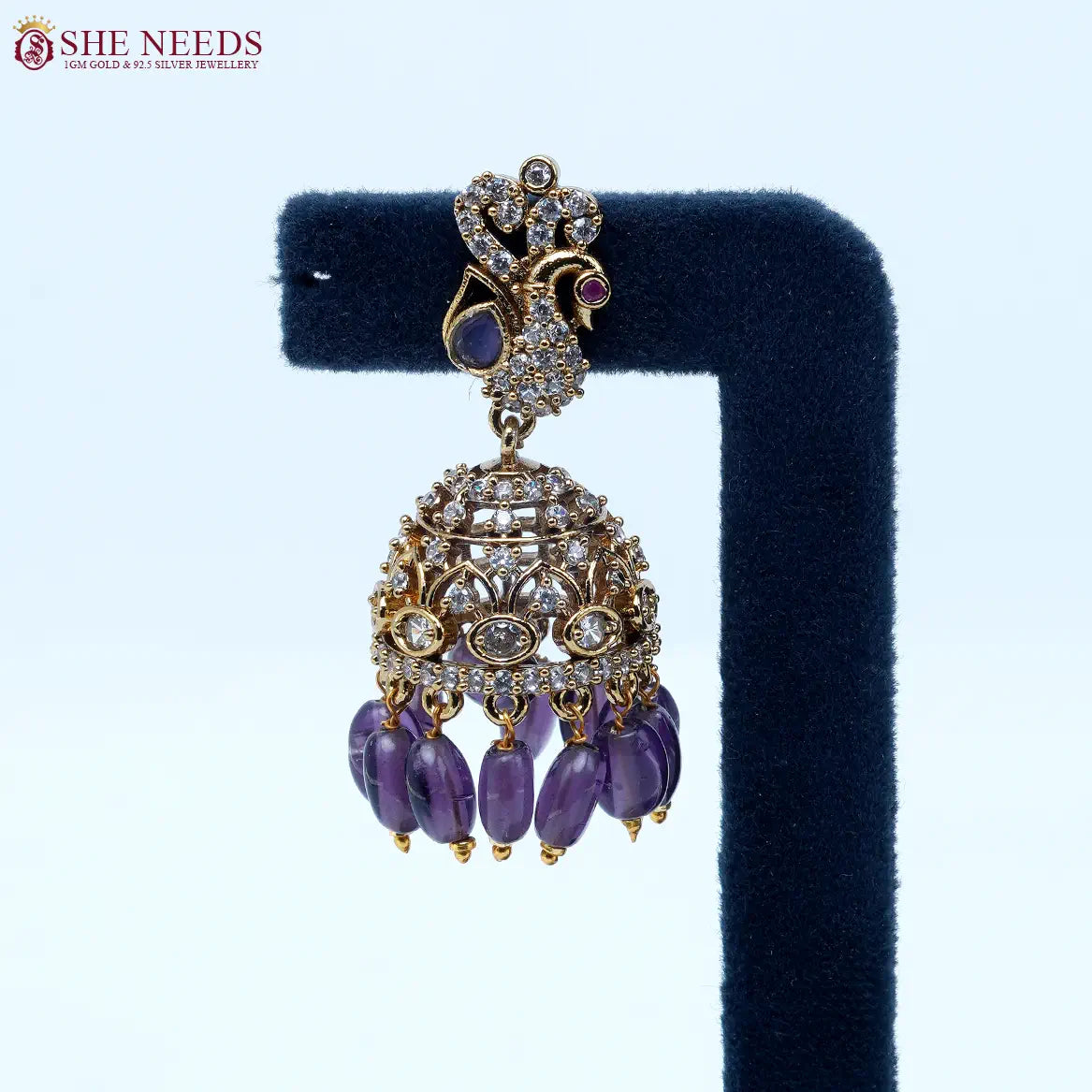 Antique gold jhumka earrings featuring crystal-studded peacock motifs, sparkling CZ stones, intricate dome detailing, and royal purple bead drops.| She Needs Jewellery