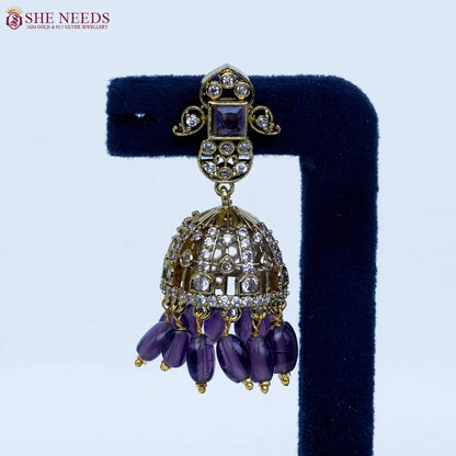 Gold-tone dome jhumka earrings featuring dazzling white stones, amethyst purple square stone accents, and rich purple bead drops in a luxurious ethnic style |She Needs Jewellery
