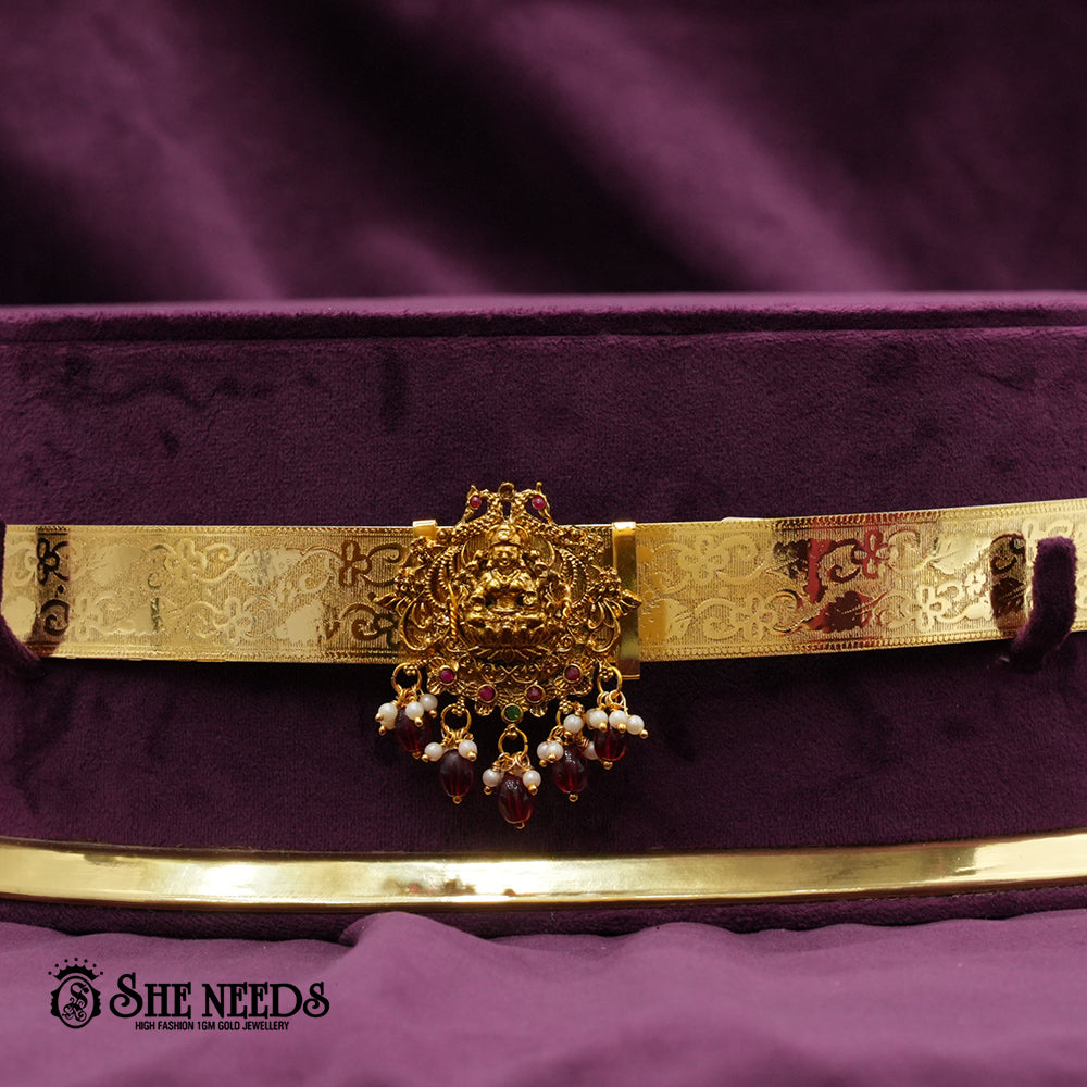 Traditional gold-finish kids vaddanam featuring a central Sri Lakshmi motif, intricate temple engravings, pearl accents, and ruby-colored bead drops.|She Needs Jewellery
