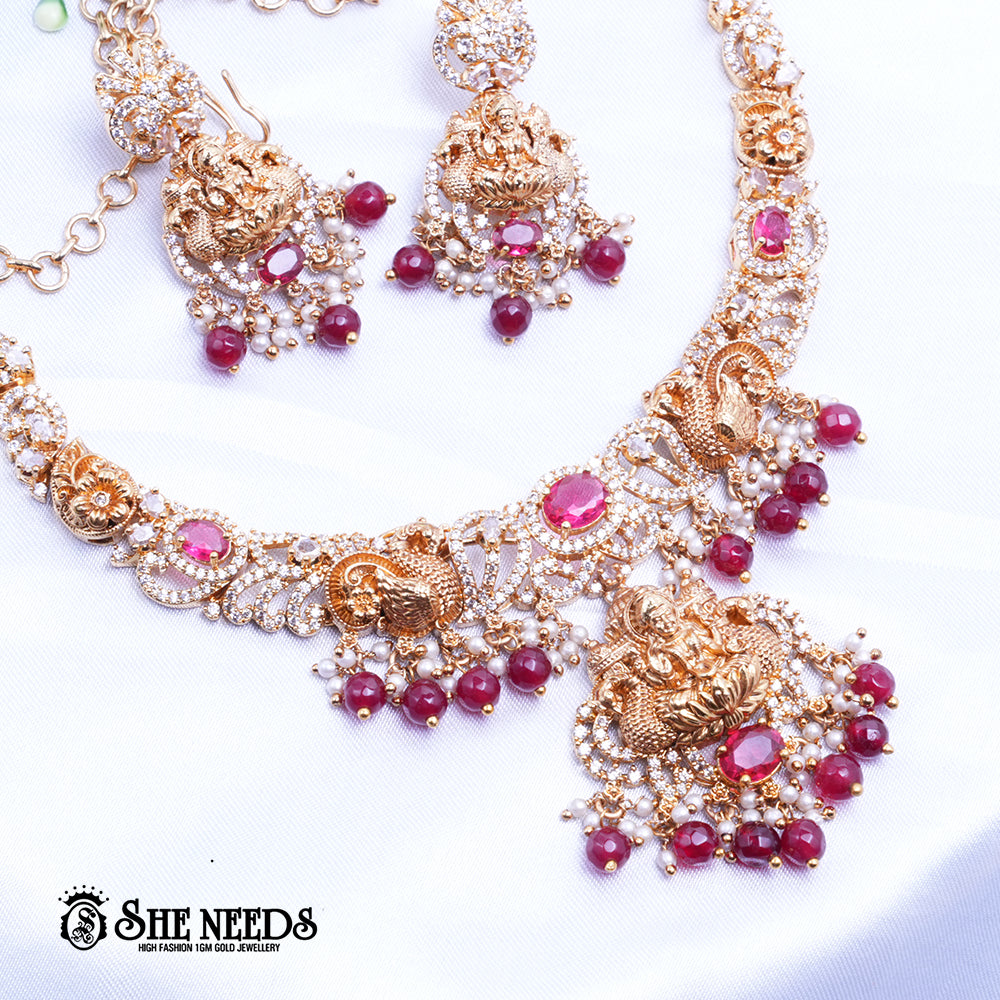 A short, high-fashion, gold-toned necklace and earring set featuring intricate carvings of a deity (likely Goddess Lakshmi) on the pendant and earrings, heavily studded with clear stones (likely CZ or white diamonds) and accented with drops of deep red ruby-colored stones/beads. The set is displayed on a white, draped cloth next to a white flower.