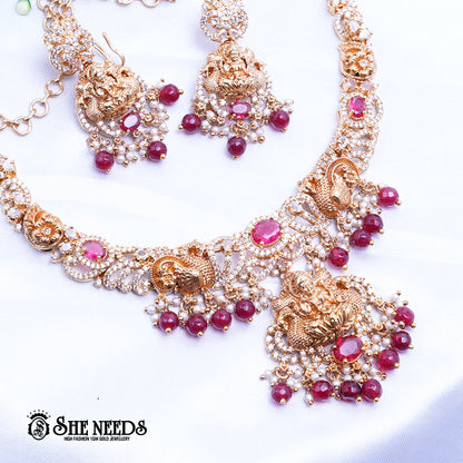 A short, high-fashion, gold-toned necklace and earring set featuring intricate carvings of a deity (likely Goddess Lakshmi) on the pendant and earrings, heavily studded with clear stones (likely CZ or white diamonds) and accented with drops of deep red ruby-colored stones/beads. The set is displayed on a white, draped cloth next to a white flower.