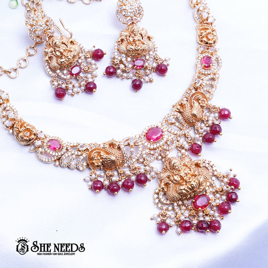 A short, high-fashion, gold-toned necklace and earring set featuring intricate carvings of a deity (likely Goddess Lakshmi) on the pendant and earrings, heavily studded with clear stones (likely CZ or white diamonds) and accented with drops of deep red ruby-colored stones/beads. The set is displayed on a white, draped cloth next to a white flower.
