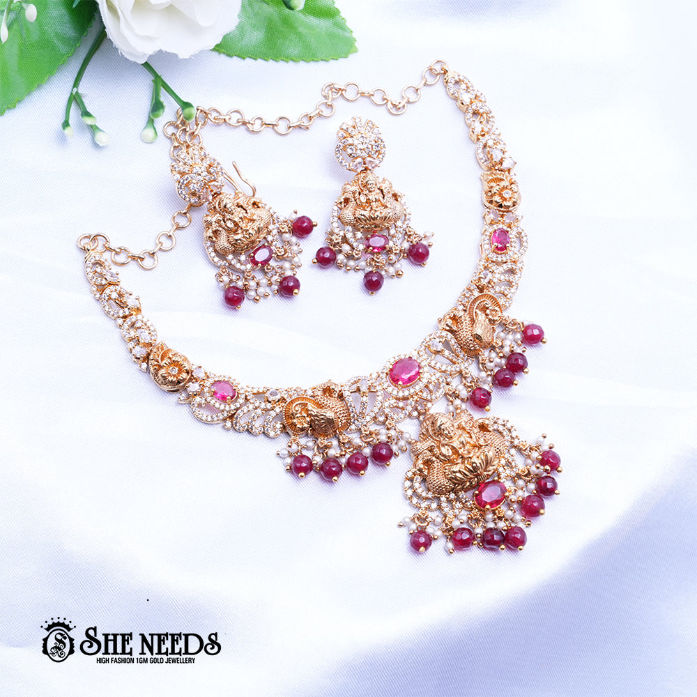 A short, high-fashion, gold-toned necklace and earring set featuring intricate carvings of a deity (likely Goddess Lakshm) on the pendant and earrings, heavily studded with clear stones (likely CZ or white diamonds) and accented with drops of deep red ruby-colored stones/beads. The set is displayed on a white, draped cloth next to a white flower.