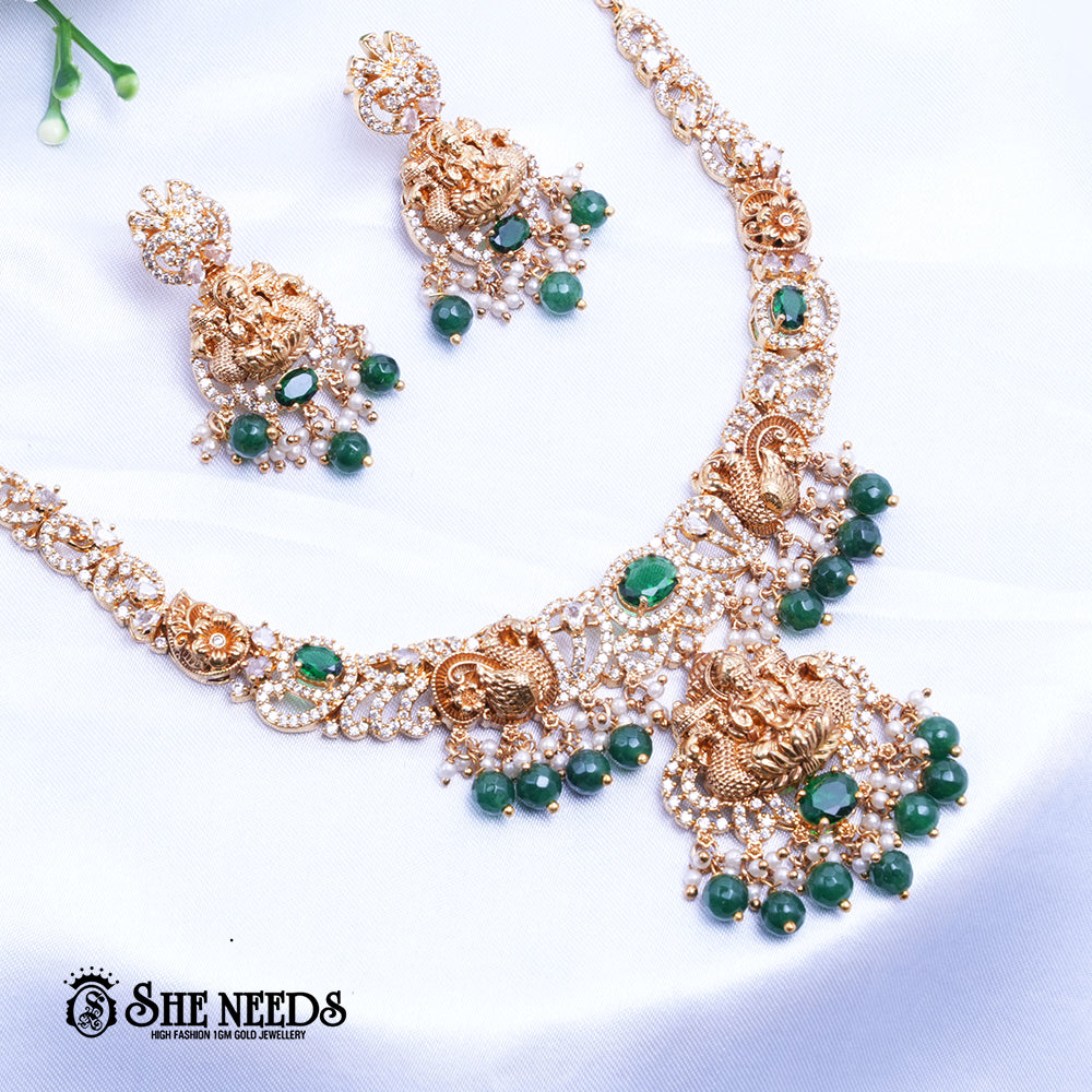 A short, high-fashion, gold-toned necklace and earring set featuring intricate carvings of a deity (likely Goddess Lakshmi) on the pendant and earrings, heavily studded with clear stones (likely CZ or white diamonds) and accented with drops of deep red ruby-colored stones/beads. The set is displayed on a white, draped cloth next to a white flower.