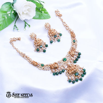 A short, high-fashion, gold-toned necklace and earring set featuring intricate carvings of a deity (likely Goddess Lakshmi ) on the pendant and earrings, heavily studded with clear stones (likely CZ or white diamonds) and accented with drops of deep red ruby-colored stones/beads. The set is displayed on a white, draped cloth next to a white flower.