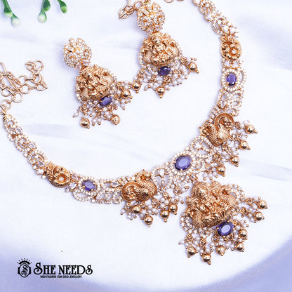 A short, high-fashion, gold-toned necklace and earring set featuring intricate carvings of a deity (likely Goddess Lakshmi) on the pendant and earrings, heavily studded with clear stones (likely CZ or white diamonds) and accented with drops of deep red ruby-colored stones/beads. The set is displayed on a white, draped cloth next to a white flower.