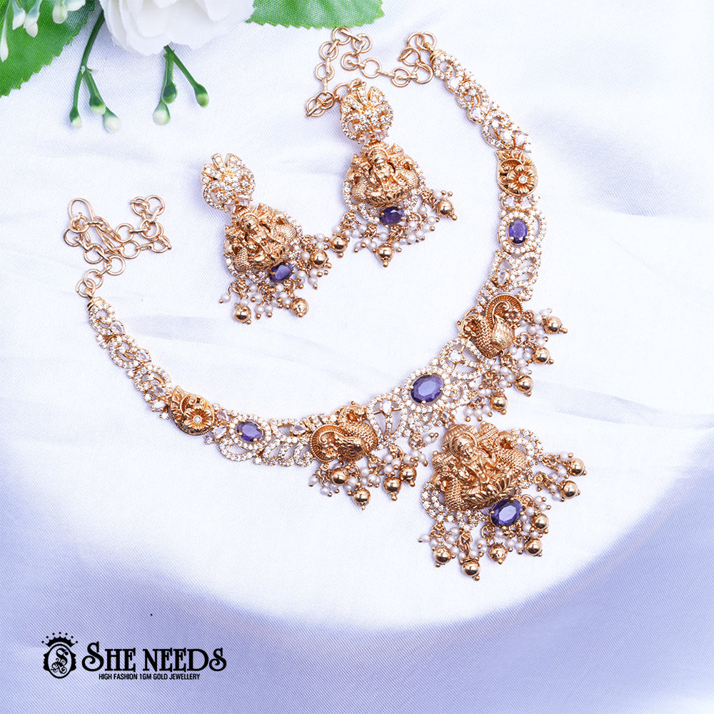 A short, high-fashion, gold-toned necklace and earring set featuring intricate carvings of a deity (likely Goddess Lakshmi ) on the pendant and earrings, heavily studded with clear stones (likely CZ or white diamonds) and accented with drops of deep red ruby-colored stones/beads. The set is displayed on a white, draped cloth next to a white flower.